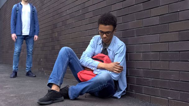 Bullied afro-american schoolboy sitting on ground, scared of offender behind - Φωτογραφία, εικόνα