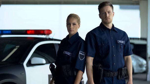 Confident woman and man police officers standing near patrol car, on duty - Foto, Imagem