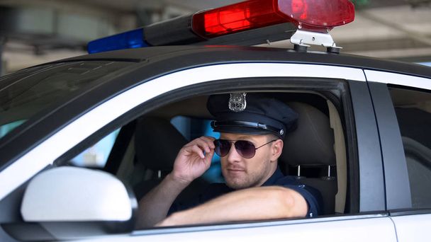 Young policeman adjusting sunglasses sitting in patrol car, responsible job - Foto, Imagem