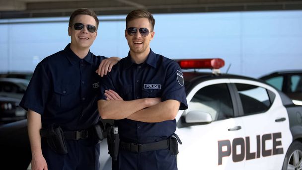 Cheerful police mates in sunglasses smiling into camera against squad car, work - Фото, изображение