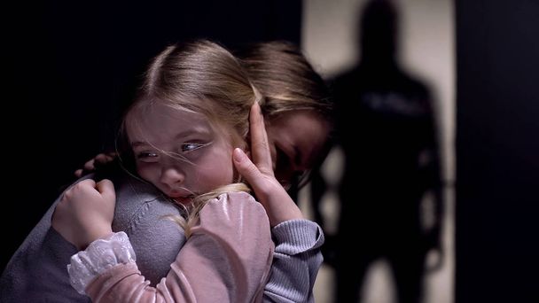 おびえた女の子は母親を抱きしめ、ドアの外に恐ろしい男性犯罪者のシルエット - 写真・画像
