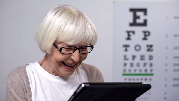 Aged lady in eyeglasses smiling and reading from tablet, focusing vision, health - Foto, afbeelding