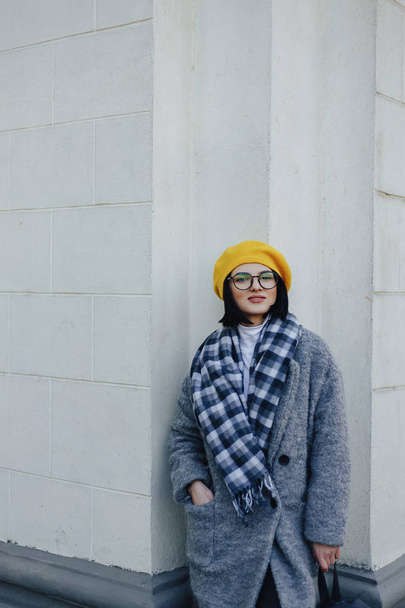 Attractive young girl in glasses in coat and yellow Beret on a simple light background - Fotó, kép
