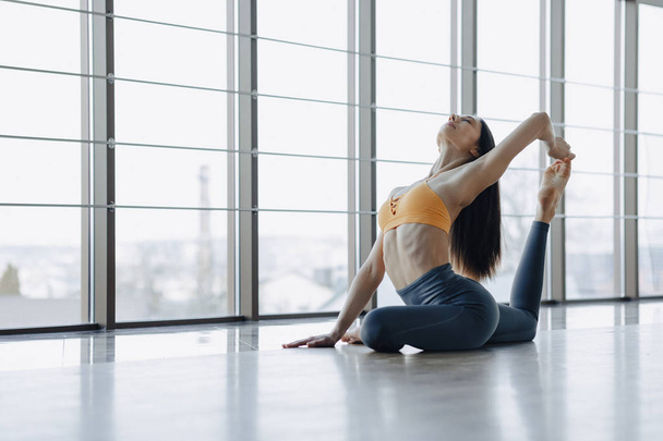 chica atractiva joven haciendo ejercicios de fitness con yoga en el suelo contra el fondo de ventanas panorámicas
 - Foto, imagen