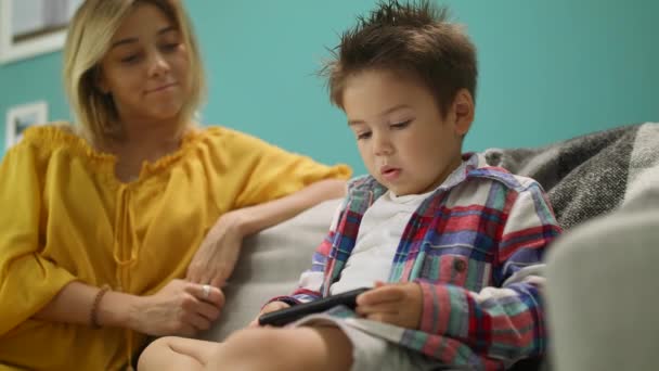 Little son with mom plays a game on a smartphone on the couch in the living room - Imágenes, Vídeo