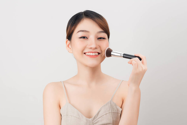Cheerful woman is doing make up on her face using brush. Beauty routine on white background. - Foto, Imagem