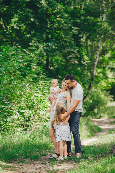 Parents and two cute little daughter in holds posing on the path in a sunny day in forest - Zdjęcie, obraz