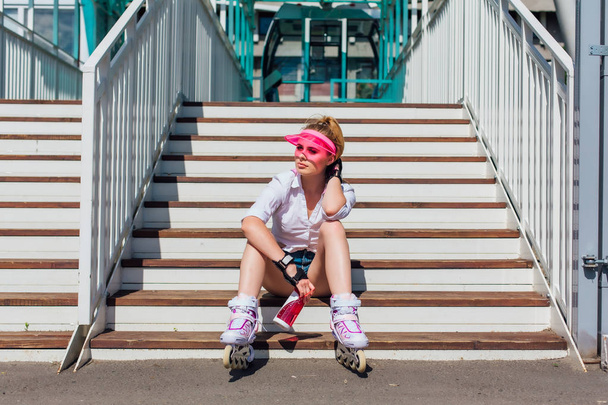 Portrait of an emotional girl in a pink cap visor wearing protective gloves and rollerblades sitting on stairs holding a bottle of water in hands - Foto, afbeelding