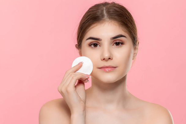 Beautiful model applyes cosmetic product to skin with white cotton sponge. Model looks at the camera isolated over pink background. - Photo, image