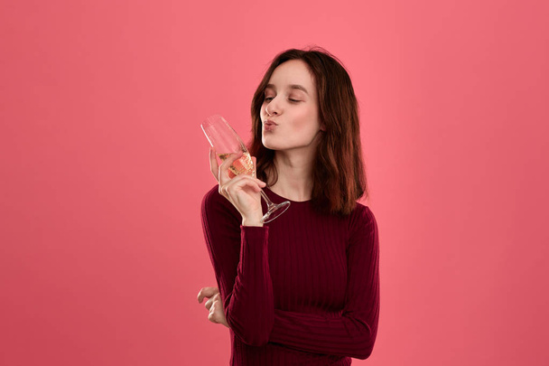 Podekscytowany Ładna Brunetka trzyma lampka szampana stojący na białym tle na ciemnym różowym tle i uśmiechnięty w aparacie. - Zdjęcie, obraz