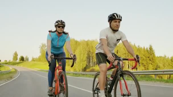 Guapo barbudo ciclista profesional masculino montando su bicicleta de carreras en la mañana junto con su novia, ambos con cascos protectores y anteojos, el sol brillando entre ellos
. - Imágenes, Vídeo