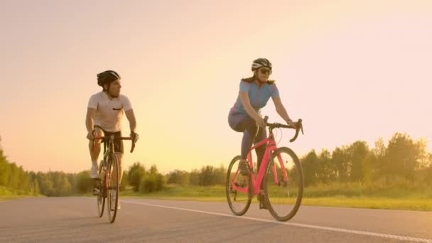 Handsome gebaarde mannelijke wielrenner zijn race-fiets in de ochtend samen met zijn vriendin, beide dragen beschermende helmen en brillen, de zon schijnt door ertussen. - Video