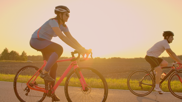 Bonito barbudo profissional ciclista masculino montando sua bicicleta de corrida na parte da manhã, juntamente com sua namorada, ambos vestindo capacetes de proteção e óculos, sol brilhando entre eles
. - Filmagem, Vídeo
