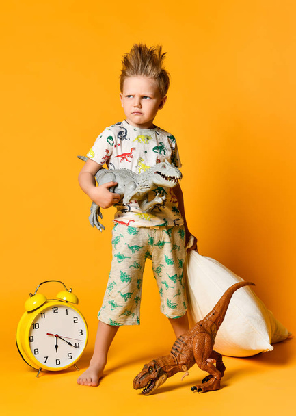 Little boy of preschool age with a pillow in his hands and a toy dinosaur waking up on an alarm clock. Photo in studio on a yellow background - Foto, Bild