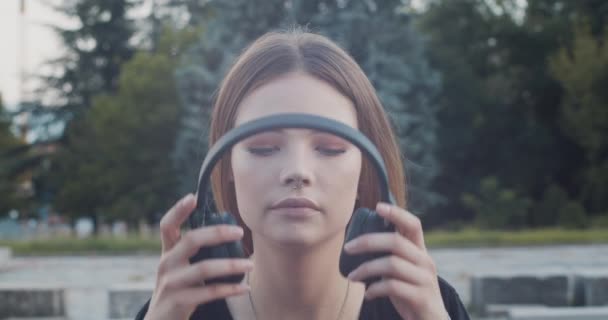 Beautiful female model listens to music on black bluetooth over the ear headphones - Imágenes, Vídeo