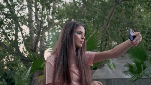 Aufnahme eines schönen Mädchens, das verträumt im alten Garten sitzt und ein Selfie mit tropischen Blättern macht - Filmmaterial, Video