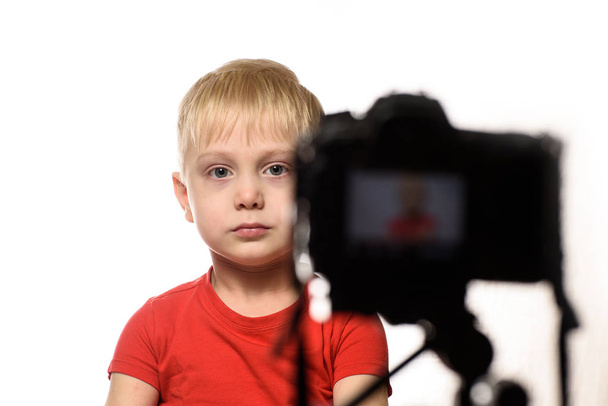 Ein ernsthafter blonder Junge nimmt ein Video mit der DSLR-Kamera auf. kleiner Videoblogger. weißer Hintergrund - Foto, Bild