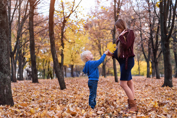 Küçük sarışın çocuk hamile anne sarı yaprak verir. Arka planda sonbahar Parkı - Fotoğraf, Görsel