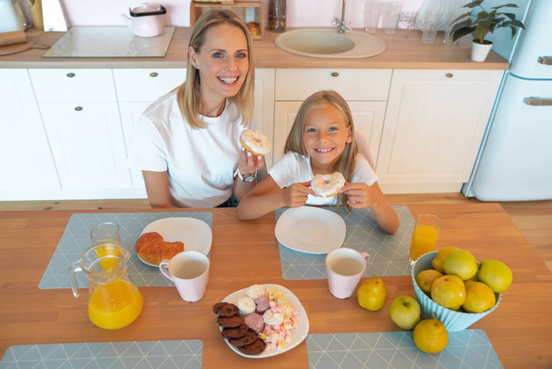 Top view mom with daughter eating delicious donuts and having fun together. dressed in white t-shirts on colored kitchen background. junk food concept - Foto, afbeelding