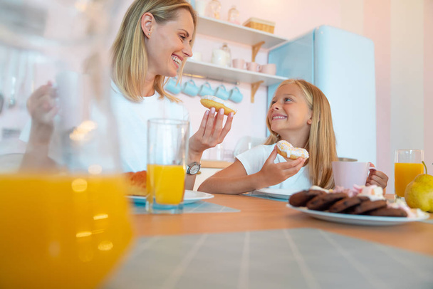 Äiti ja hänen tyttärensä syövät herkullisia donitseja ja pitävät hauskaa yhdessä. pukeutunut valkoinen t-paitoja värillinen keittiö tausta
. - Valokuva, kuva
