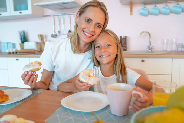 Nettes Foto von Mutter und Tochter beim Frühstück mit Donuts und Tee. Kuscheln und in die Kamera schauen - Foto, Bild