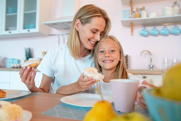 Nettes Foto von Mutter und Tochter beim Frühstück mit Donuts und Tee. Kuscheln und in die Kamera schauen - Foto, Bild