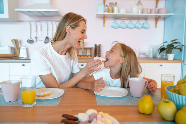 moeder en dochter zijn gek rond in de keuken met donuts. leuke familiefoto. gekleed in witte tshirts - Foto, afbeelding