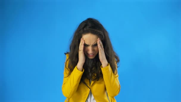 Beautiful girl is suffering from a migraine standing on a blue background, slow motion - Footage, Video