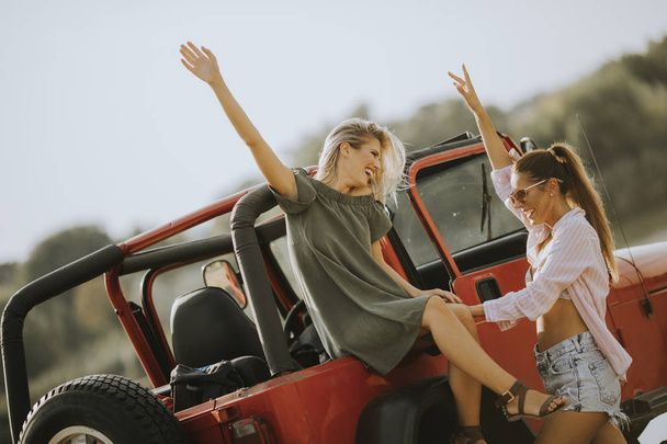 Duas jovens mulheres se divertindo no capô de um carro junto ao lago no dia quente de verão
 - Foto, Imagem