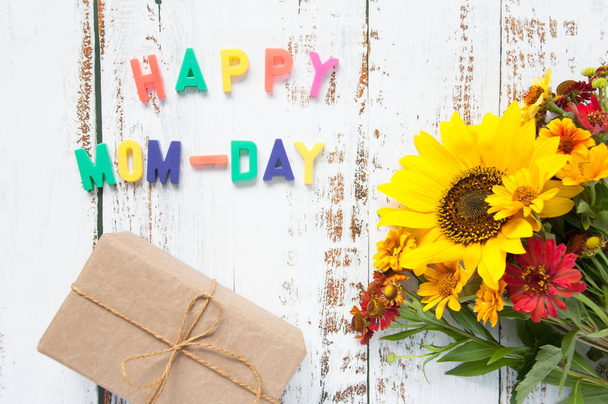 Happy mother greeting card with flowers bouquet with sunflower with a gift on a light wooden background with bright letters text congratulation. Happy mom-day. - Фото, изображение