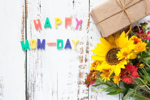 Happy mother greeting card with flowers bouquet with sunflower with a gift on a light wooden background with bright letters text congratulation. Happy mom-day. - Фото, изображение