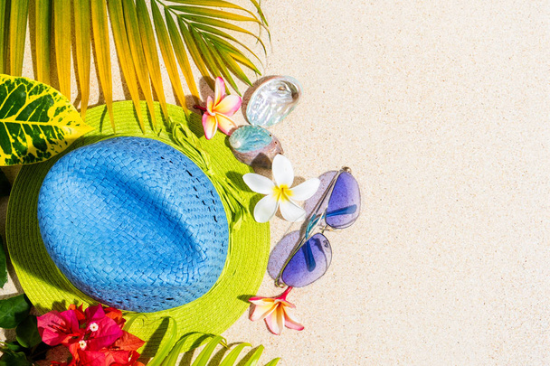 Blue and green straw hat with sunglasses, sea shells and frangipani flowers with green palm leafs on sand, summer vacation concept, top view, copy sapce - Photo, Image