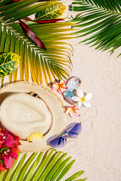 青いサングラス、カラフルな海の貝殻、砂の上にフランジパニの花と緑のヤシの葉、夏休みの概念、垂直構成とベージュのわら帽子  - 写真・画像