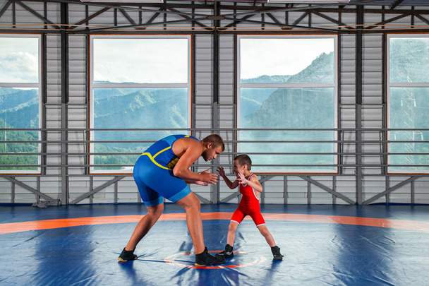 スポーツタイツの小さなレスラーの少年は、ジムのレスリングカーペットの上で大人の男性レスラーと格闘します。子どもの力と武道の訓練の概念。子供たちにグレコローマレスリングを教える - 写真・画像