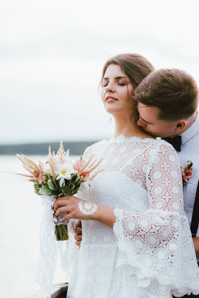 Happy kissing newly married couple, smiling bride brunette young woman with boho style bouquet with groom, outdoors - Zdjęcie, obraz