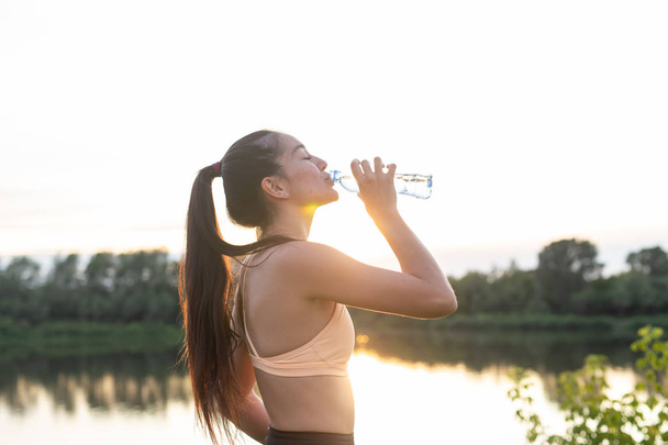 Vista lateral de la mujer fitness beber agua al atardecer
 - Foto, imagen