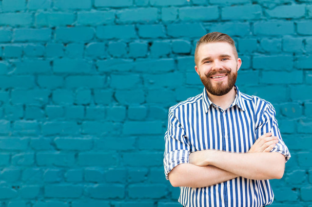 Portrait of a young positive stylish smiling guy with a mustache and beard against the background of a blue brick wall with copyspace. Vacation concept in the city. - Photo, Image