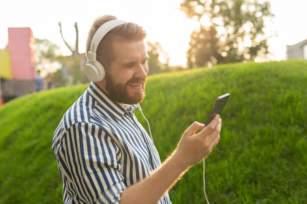 Pozytywny młody męski Hipster słucha ulubionego radia internetowego za pomocą słuchawek smartphone i szybkiego bezprzewodowego Internetu w parku w słoneczny ciepły letni dzień. - Zdjęcie, obraz