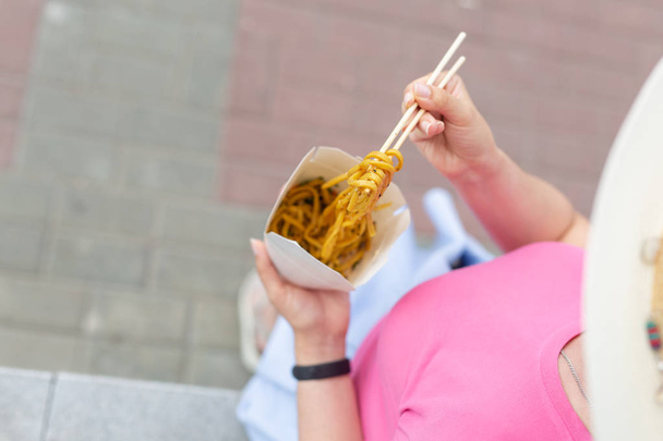Beautiful young unidentified woman in a hat eats Chinese noodles while walking in the park on a warm summer day. Takeaway food concept. - Photo, image