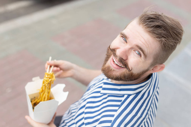 Young male hipster with a mustache and beard is eating chinese noodles with wooden chopsticks from a white lunch box. Asian cuisine concept. - Photo, Image