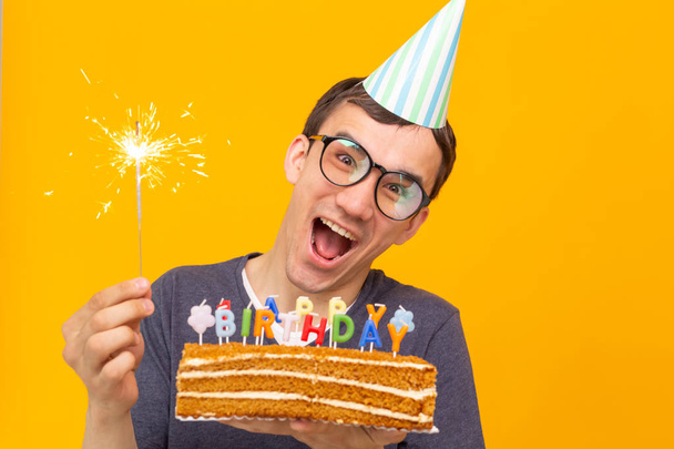 Gözlük ve kağıt tebrik şapkadeli neşeli genç adam sarı bir arka plan üzerinde duran kek mutlu doğum günü tutan. Jubilee tebrikler kavramı. - Fotoğraf, Görsel