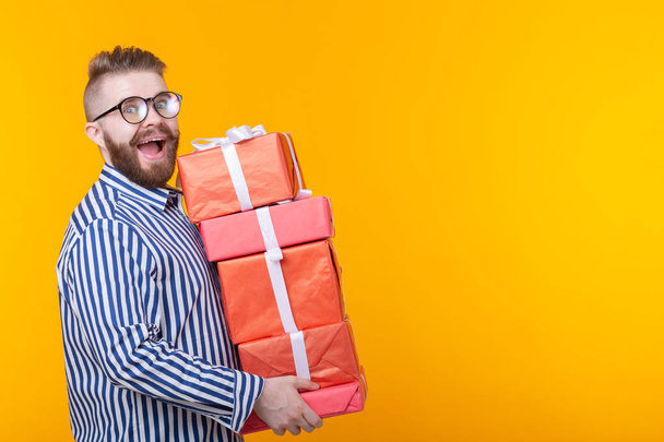 Surprised young hipster guy in glasses holding a large stack of gift boxes on a yellow background with copy space. The concept of New Year and Christmas gifts - 写真・画像