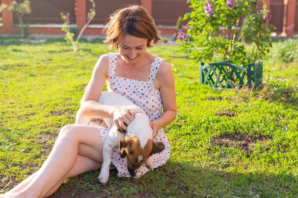 Positive cheerful woman playing with her beloved dog Jack Russell Terrier in the yard of her country house on a sunny summer day. Concept of love for animals and family recreation. - Photo, Image