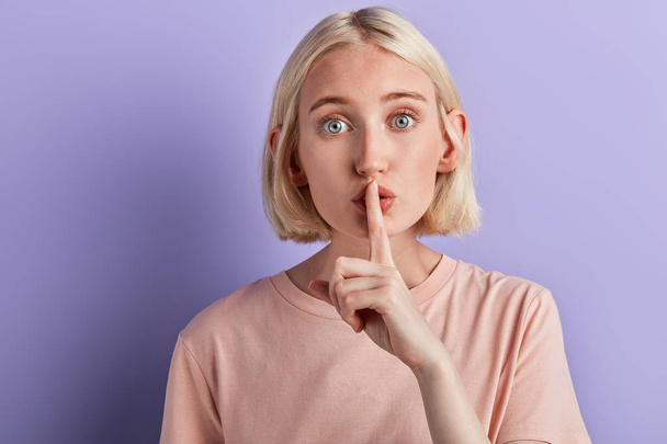 blonde girl showing shhh sign with finger to lips over violet background - 写真・画像