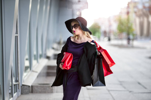 Stylish blonde woman in black coat, sunglasses and hat with shopping bags walks along the street - Foto, Bild