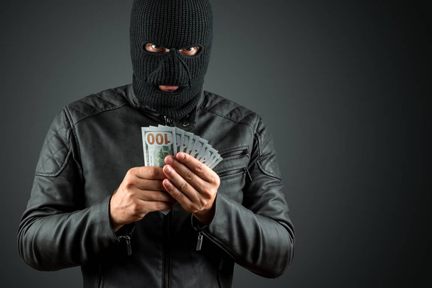 Einbrecher, ein Schläger mit Sturmhaube hält vor dunklem Hintergrund Dollars in den Händen. Raub, Hacker, Verbrechen, Diebstahl. Kopierraum. - Foto, Bild