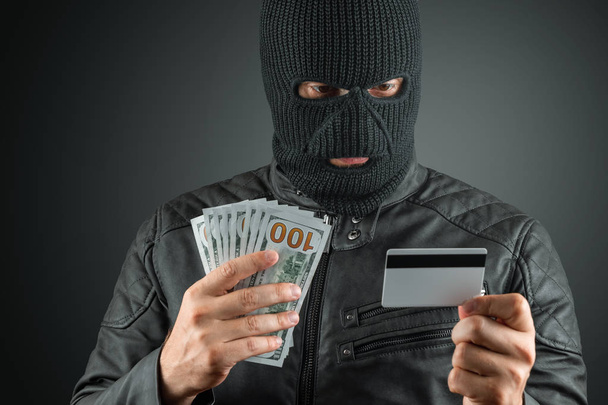 Robber, a thug in a balaclava holds a credit card in his hands on a dark background. Robbery, hacker, crime, theft. Copy space. - Foto, Bild