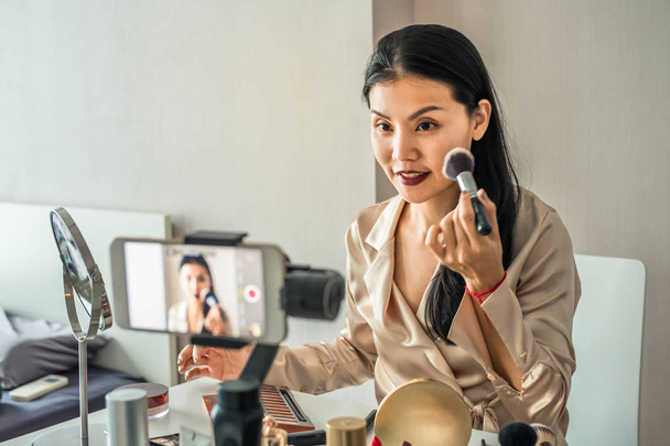 Jolie asiatique femme enregistrer une vidéo avec téléphone intelligent tout en utilisant maquillage pour cours en ligne. Tutoriel beauté
 - Photo, image