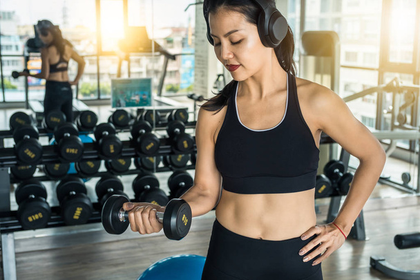 Mujer asiática entrenando brazos en el gimnasio con pesas mientras escucha música con auriculares
 - Foto, imagen