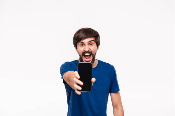 Hombre morena bien parecido en una camisa azul con barba y bigotes mostrando la pantalla del teléfono inteligente en la cámara de pie aislado sobre fondo blanco
. - Foto, imagen